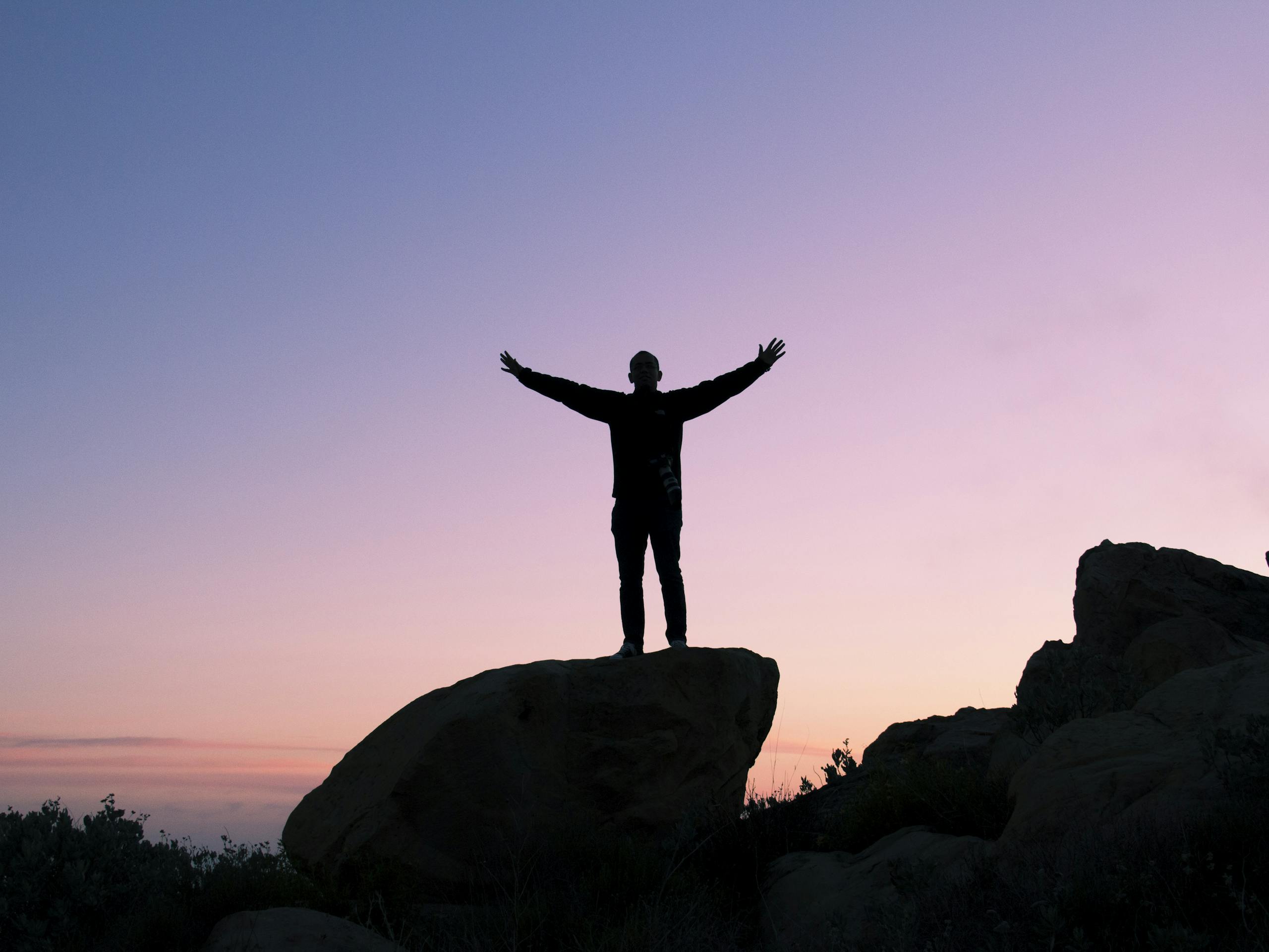 Silhouette of a man on a rock with arms raised, embracing the sunset sky.